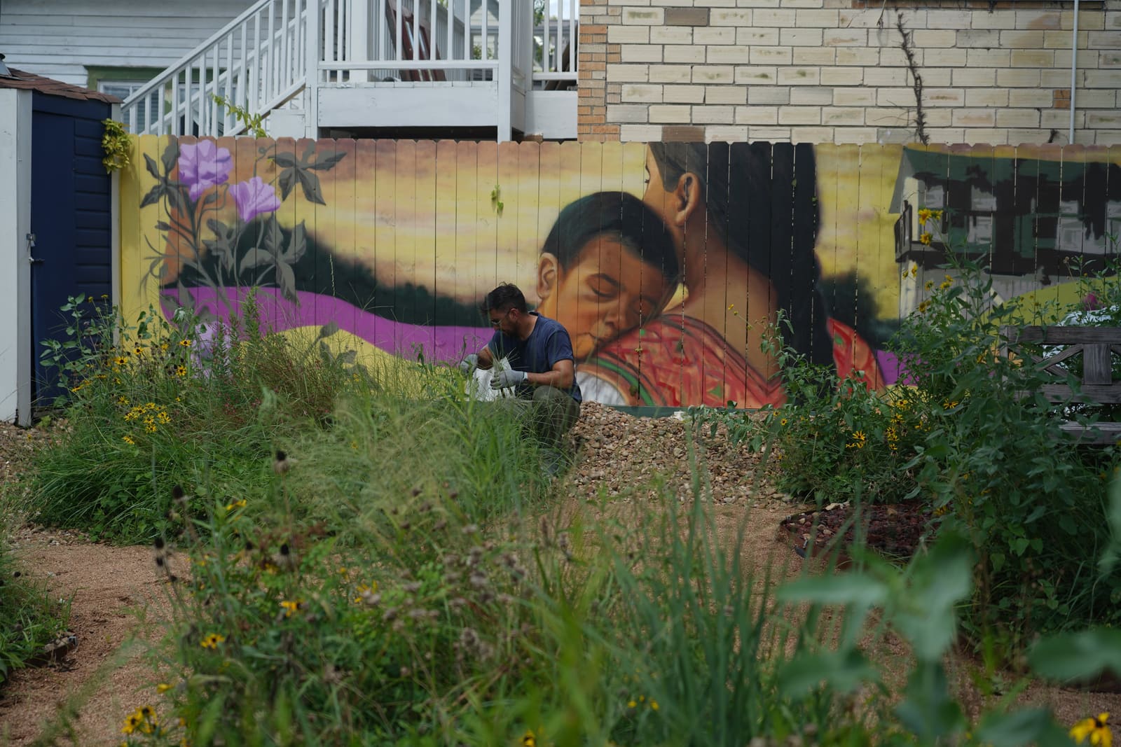 A painted fence mural of a mother holding a sleeping child behind a garden bed